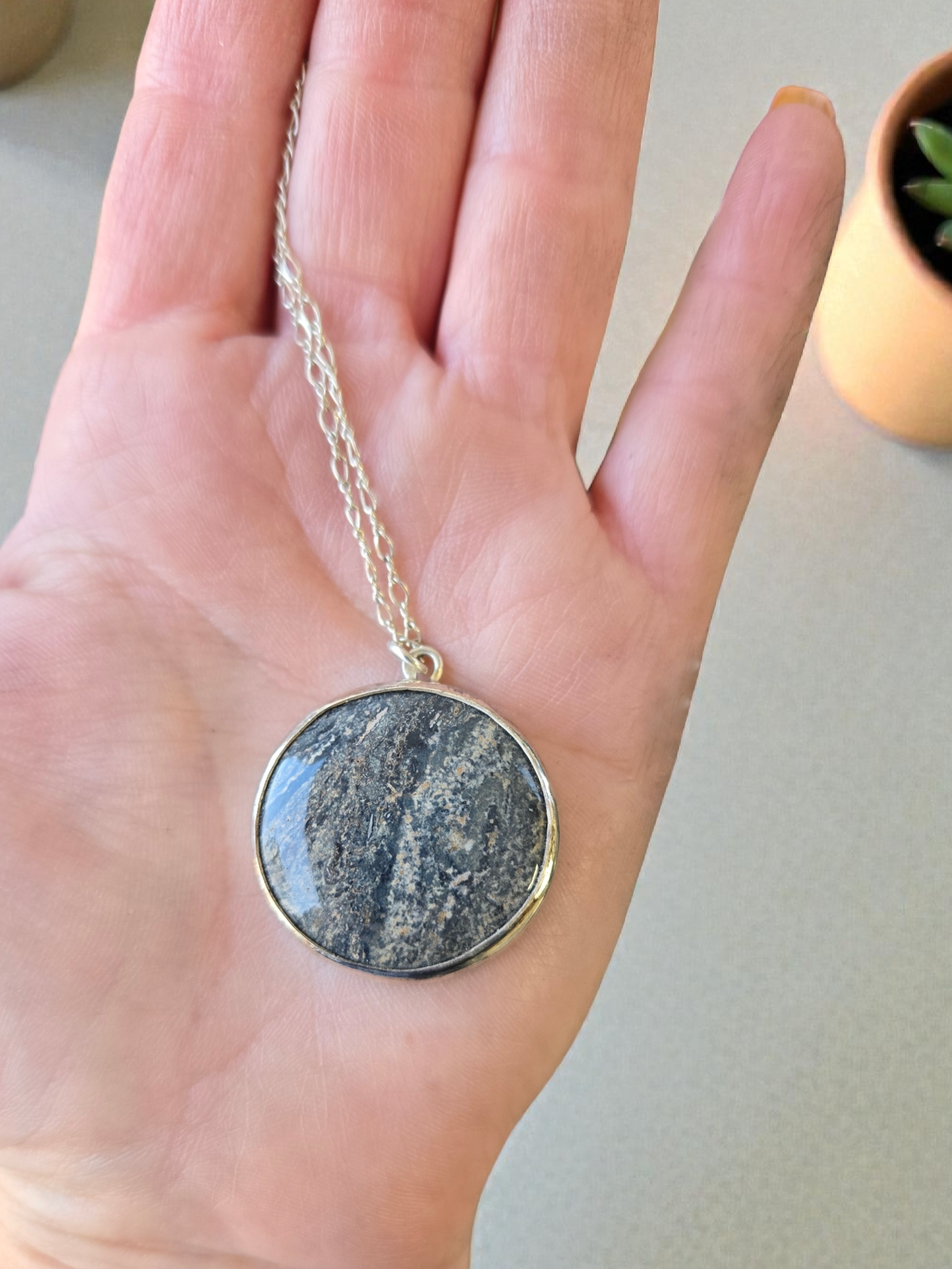 Blue tan round cabochon in a sterling bezel on a small cable chain shown in maker's hand.